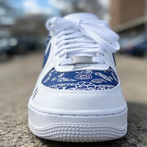 May include: White and blue athletic shoe with a paisley design on the toe box. The shoe features white laces, a silver "AIR" tag, and a white sole. The shoe has blue accents and a classic sneaker design.