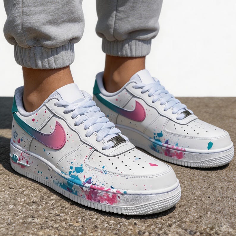 May include: White leather sneakers with a colorful paint splatter design. The shoes feature a pink and blue gradient swoosh and accents, with matching paint splatters in pink and blue. The laces are white, and the soles are white.