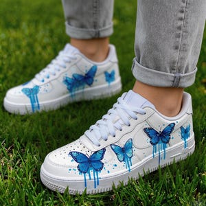Op de afbeelding: Witte sneakers met blauwe vlinderontwerpen. De schoenen hebben een witte basis, blauwe vlinderillustraties en druipende verfeffecten. Witte veters en een witte zool met grijze rand. Op een groene grasachtergrond.