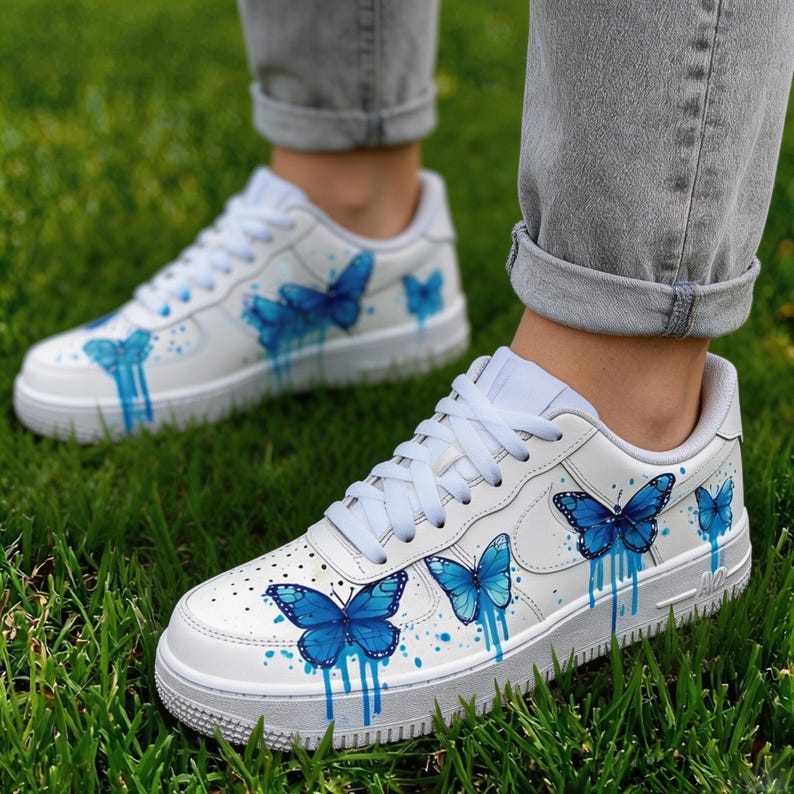 Op de afbeelding: Witte sneakers met blauwe vlinderontwerpen. De schoenen hebben een witte basis, blauwe vlinderillustraties en druipende verfeffecten. Witte veters en een witte zool met grijze rand. Op een groene grasachtergrond.
