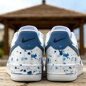 May include: Pair of white and blue Nike Air Force 1 sneakers. The shoes feature a white base with blue paint splatter details and blue accents on the heel and swoosh logo. The soles are white with blue detailing.