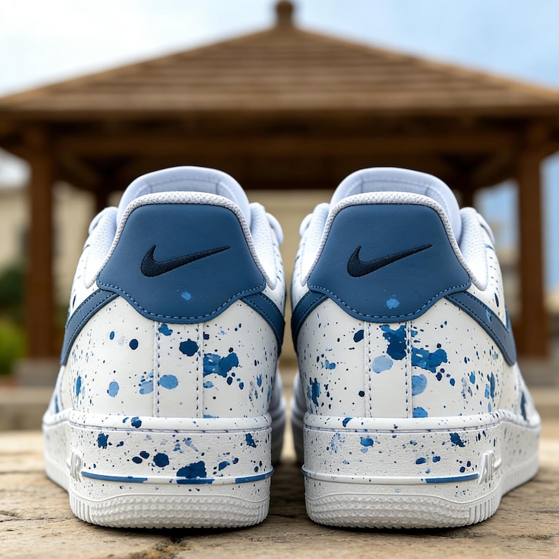 May include: Pair of white and blue Nike Air Force 1 sneakers. The shoes feature a white base with blue paint splatter details and blue accents on the heel and swoosh logo. The soles are white with blue detailing.