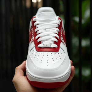 Puede incluir: Zapatilla deportiva blanca y roja con cordones blancos. La zapatilla tiene un acento rojo en el tal&oacute;n y la parte superior, con una suela blanca y una puntera perforada. La zapatilla est&aacute; sujeta por una mano.
