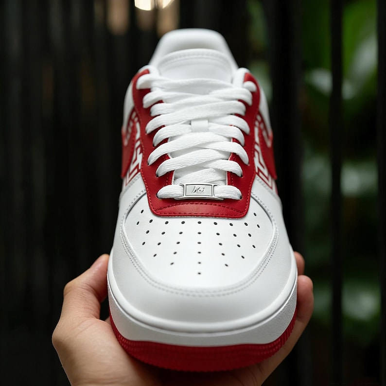 Puede incluir: Zapatilla deportiva blanca y roja con cordones blancos. La zapatilla tiene un acento rojo en el tal&oacute;n y la parte superior, con una suela blanca y una puntera perforada. La zapatilla est&aacute; sujeta por una mano.