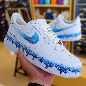 May include: White sneakers with a blue gradient design. The Nike swoosh and the bottom edge of the shoe feature a dripping blue paint effect. The word "hoebe" is written in blue script on the side.