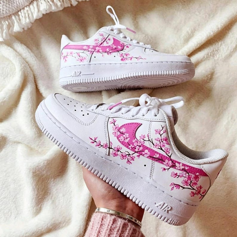 May include: White sneakers with pink floral design. The shoes feature a pink swoosh and pink cherry blossom branches. The soles are white with the word "AIR" printed on the side. The laces are white.