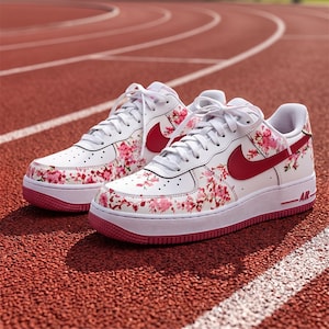 May include: White trainers with a floral pattern and red accents. The shoes feature a white base with pink floral designs, a red swoosh, and a pink sole. The laces are white, and the trainers are on a red track.