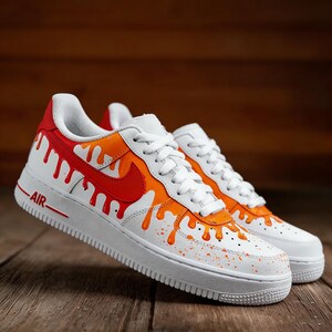 May include: White sneakers with orange and red paint drip design. The shoes have white laces, a white sole, and the word "AIR" in red. The Nike swoosh is red. The design is on a wooden surface.