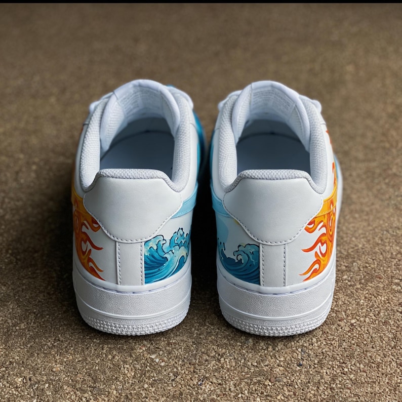 May include: Pair of white sneakers with custom artwork. One shoe features a fiery sun design in orange and yellow, while the other showcases blue ocean waves. The shoes have a white sole and accents.