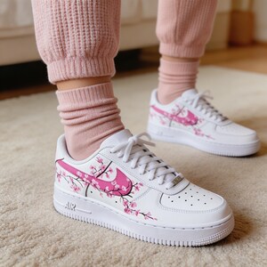 May include: White sneakers with pink floral designs and a pink Nike swoosh. The shoes have white laces and a white sole. The sneakers are on a light-colored carpet.