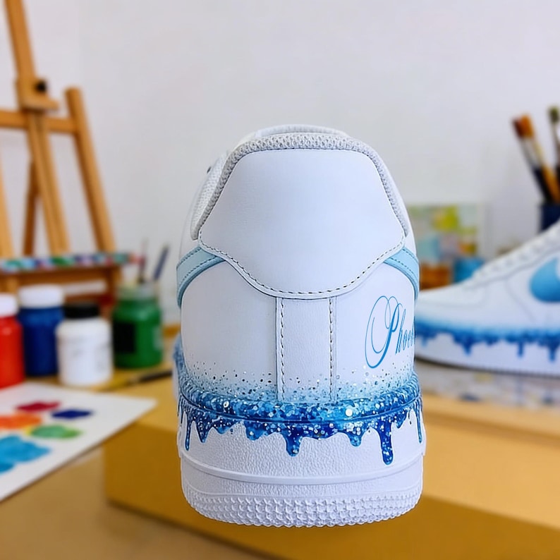 May include: White and light blue custom sneaker with a blue glitter and drip design. The word "Phoenix" is written in cursive on the heel. The shoe has a white sole and light blue accents.