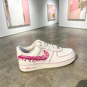 May include: White leather sneaker with pink accents. The shoe features a pink swoosh design with a dripping effect and pink trim. The word "AIR" is visible on the sole. The sneaker is displayed in an art gallery.
