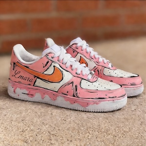May include: A pair of custom pink and white sneakers with orange accents. The shoes feature a unique design with black outlines, white laces, and the name "Lmara" on the side. The soles are white with a pink drip effect.