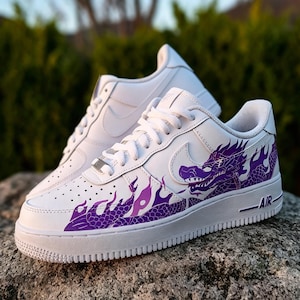 May include: White sneakers with a purple dragon design on the side. The shoes have white laces, a white sole, and the word "AIR" printed in black. The dragon design is a prominent feature, adding a unique aesthetic to the footwear.