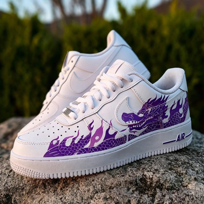 May include: White sneakers with a purple dragon design on the side. The shoes have white laces, a white sole, and the word "AIR" printed in black. The dragon design is a prominent feature, adding a unique aesthetic to the footwear.