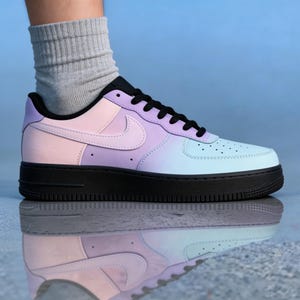 Op de afbeelding: Een pastelkleurige sneaker met een zwarte zool en veters. De schoen heeft een gradi&euml;ntontwerp, dat overgaat van roze naar paars naar lichtblauw. Het iconische swoosh-logo is zichtbaar aan de zijkant.