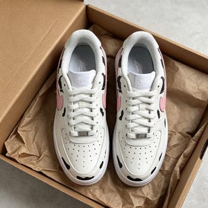 Op de afbeelding: Paar witte leren sneakers met een koeienprint. De schoenen hebben zwarte vlekken, roze accenten en witte veters. De sneakers zitten in een kartonnen doos met bruin papier.