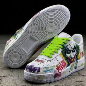 May include: White sneakers with a Joker-themed design. The shoes feature a graphic of the Joker's face, neon green laces, and colorful graffiti-style accents. The word "FAIER" is visible on the toe.