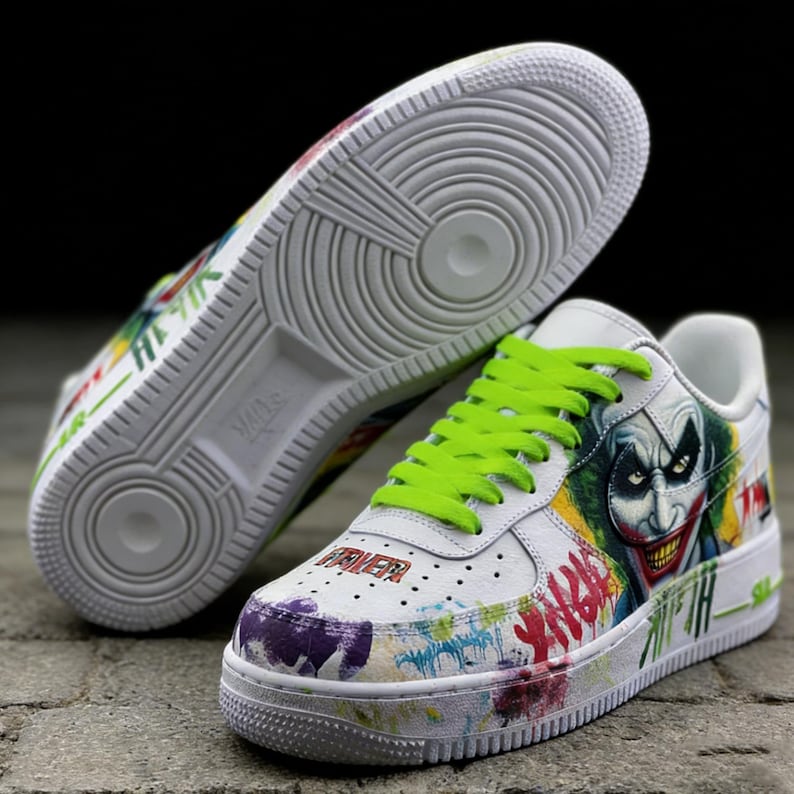 May include: White sneakers with a Joker-themed design. The shoes feature a graphic of the Joker's face, neon green laces, and colorful graffiti-style accents. The word "FAIER" is visible on the toe.