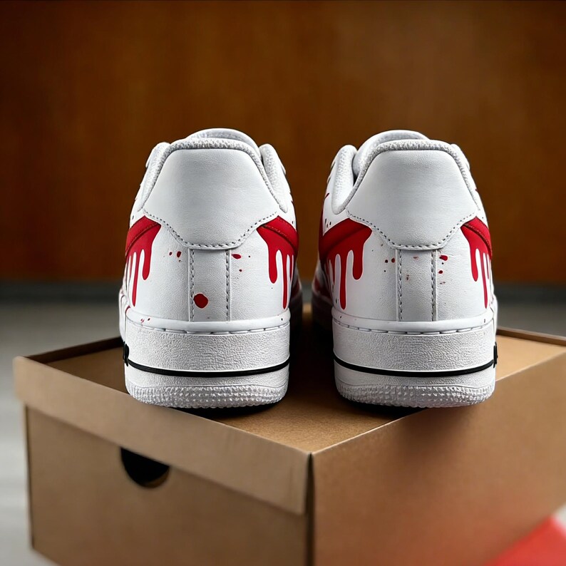 Op de afbeelding: Paar witte sneakers met een rood verfspatontwerp op de achterkant. De schoenen hebben een witte zool met een zwarte streep en staan op een bruine kartonnen doos.