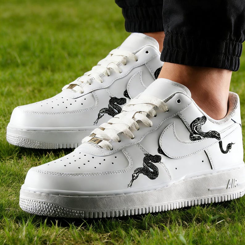 May include: White leather sneakers with black snake graphics. The shoes feature a classic design with a low-top silhouette, white laces, and a textured rubber sole. The Nike swoosh is visible on the side.