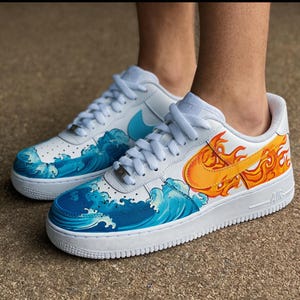 May include: White sneakers with custom wave and flame designs. The shoes feature blue ocean waves on the toe and side, and orange and yellow flames on the side. The Nike swoosh is visible in blue and orange.
