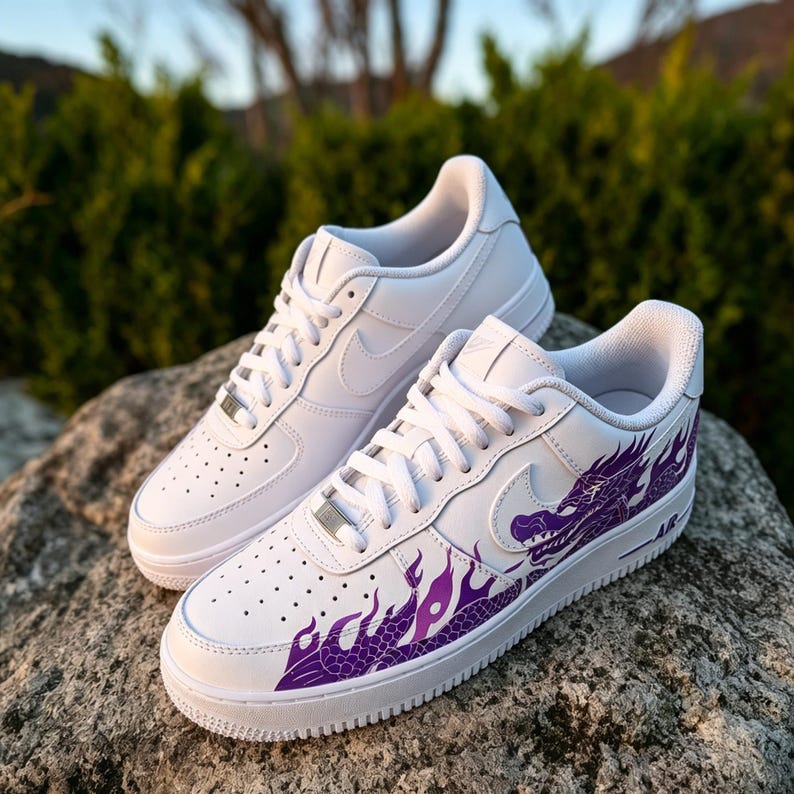 May include: Pair of white sneakers with a purple dragon design on the side. The shoes have white laces, a white sole, and the Nike swoosh logo. The sneakers are sitting on a gray rock.