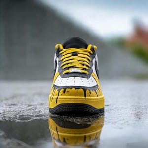 Puede incluir: Primer plano de una zapatilla deportiva amarilla, negra y blanca. El zapato presenta una suela amarilla, detalles negros y una parte superior blanca con detalles de goteo de pintura negra. Los cordones amarillos y la leng&uuml;eta negra completan el dise&ntilde;o. El zapato se refleja en una superficie mojada.