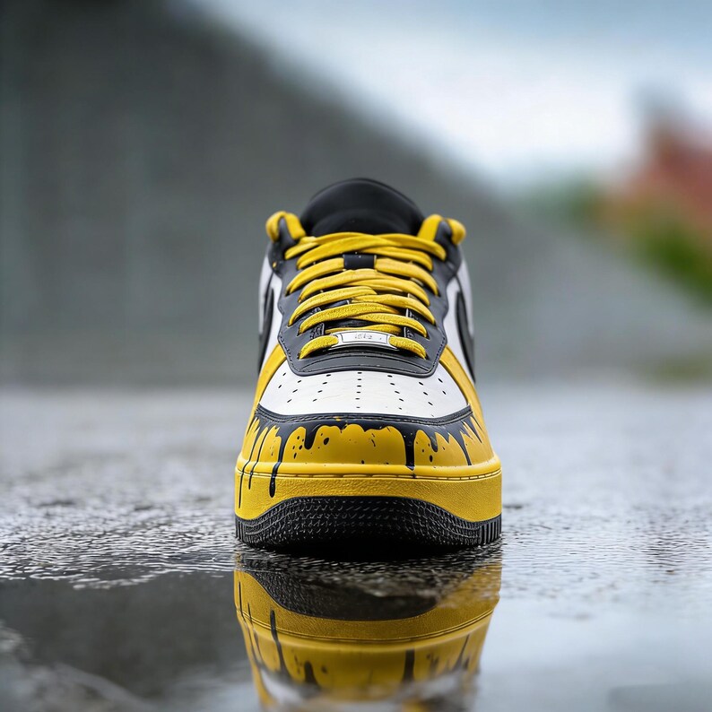 Puede incluir: Primer plano de una zapatilla deportiva amarilla, negra y blanca. El zapato presenta una suela amarilla, detalles negros y una parte superior blanca con detalles de goteo de pintura negra. Los cordones amarillos y la leng&uuml;eta negra completan el dise&ntilde;o. El zapato se refleja en una superficie mojada.