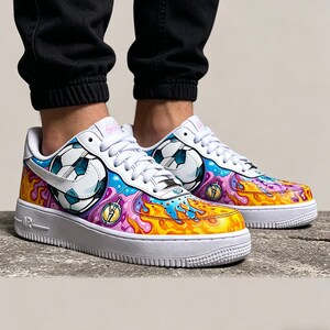 May include: White sneakers with a soccer ball design and colorful graffiti-style accents. The shoes feature a white sole and laces, with a vibrant mix of blue, yellow, pink, and purple hues. The design includes a soccer ball graphic on the side.
