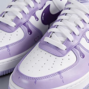 May include: Close-up of white and lavender sneakers with white laces. The sneakers feature a purple Nike swoosh and a dripping paint design in lavender. The toe box has small ventilation holes.