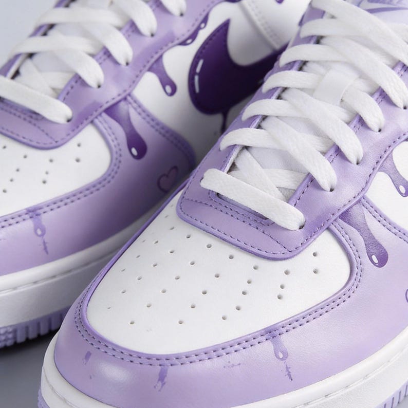 May include: Close-up of white and lavender sneakers with white laces. The sneakers feature a purple Nike swoosh and a dripping paint design in lavender. The toe box has small ventilation holes.