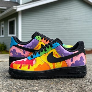 May include: A pair of custom-painted sneakers with a vibrant, colorful design. The shoes feature a black sole and laces, with a rainbow of colors splashed across the upper, including yellow, red, purple, and blue. A black Nike swoosh is visible on the side.
