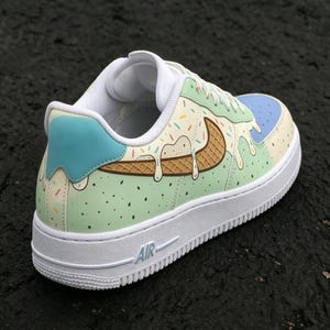 May include: A white-soled sneaker with a custom design featuring an ice cream cone theme. The shoe has a light green base with a melting ice cream design, a blue heel tab, and a brown waffle cone-shaped swoosh.