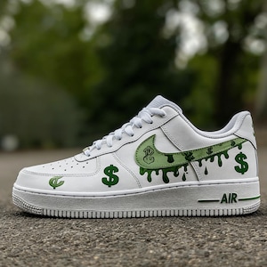May include: White low-top sneaker with green dollar sign and dripping paint design. The Nike swoosh is green with a dollar sign graphic. The sole is white with green accents and the word "AIR".