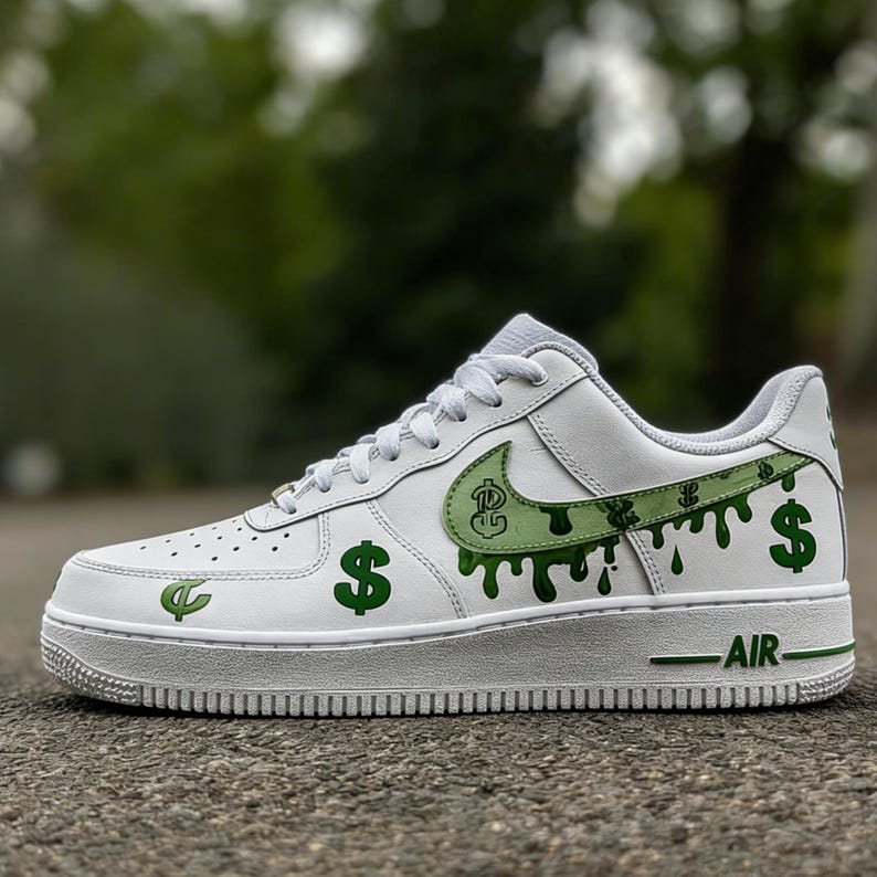 May include: White low-top trainer with green dollar sign and dripping paint design. The Nike swoosh is green with a dollar sign graphic. The sole is white with green accents and the word "AIR".
