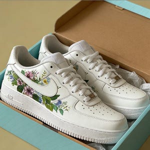 May include: White trainers with floral patterns, presented in a light blue box. The trainers have a white base, adorned with colorful flowers and green leaves. The Nike swoosh is visible. The box is light blue with a brown interior.