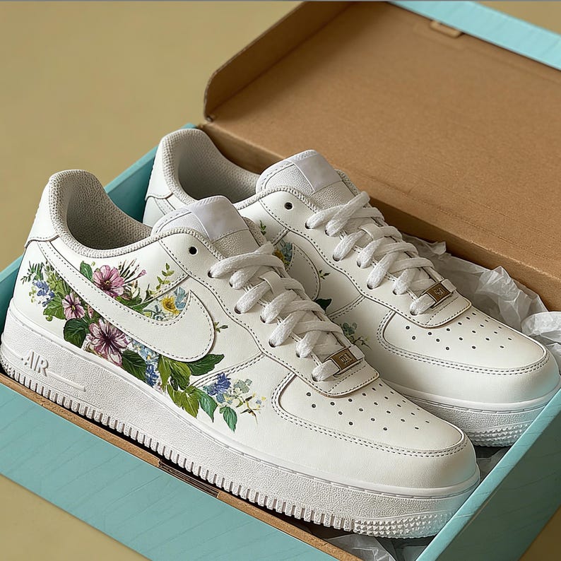 May include: White trainers with floral patterns, presented in a light blue box. The trainers have a white base, adorned with colorful flowers and green leaves. The Nike swoosh is visible. The box is light blue with a brown interior.