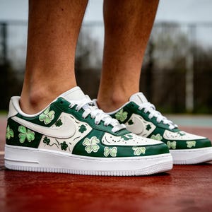May include: White and green sneakers with a shamrock design. The shoes feature a white sole, white laces, and a green and white upper with shamrock graphics. The Nike swoosh is visible on the side.