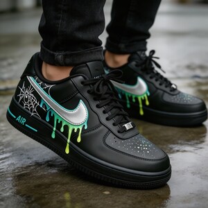 May include: Black sneakers with a silver swoosh and dripping green and yellow paint design. The shoes have a white spiderweb graphic and the word "AIR" in white. The laces are black.