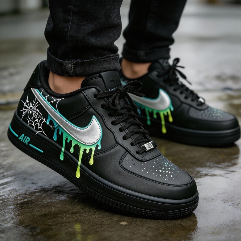 May include: Black sneakers with a silver swoosh and dripping green and yellow paint design. The shoes have a white spiderweb graphic and the word "AIR" in white. The laces are black.
