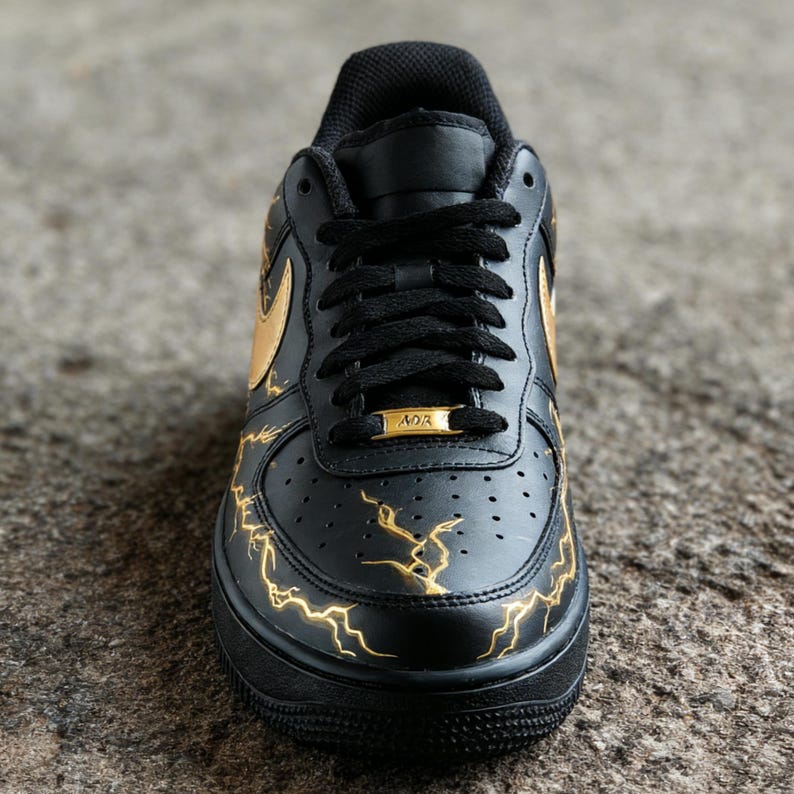 Op de afbeelding: Zwarte en gouden sportschoen met een bliksemontwerp. De schoen heeft een zwarte basis met gouden accenten, waaronder de Nike swoosh en bliksemgraphics. De veters zijn zwart en de zool is zwart.