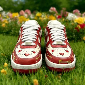 May include: A pair of red and white sneakers with heart and arrow designs. The sneakers have white laces, a white sole, and are set against a backdrop of green grass and colorful flowers. The design includes red hearts and gold arrows.