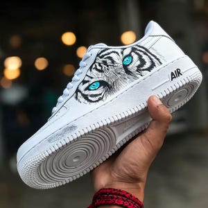 May include: White athletic shoe with a tiger eye design. The shoe features a detailed black and white tiger face with bright blue eyes. The word "AIR" is printed on the side of the shoe.