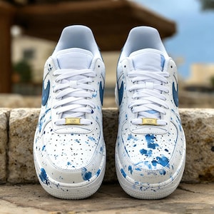 May include: Pair of white sneakers with blue paint splatter design. The shoes have white laces, blue swoosh logos, and gold-colored lace locks. The sneakers are displayed on a stone surface.