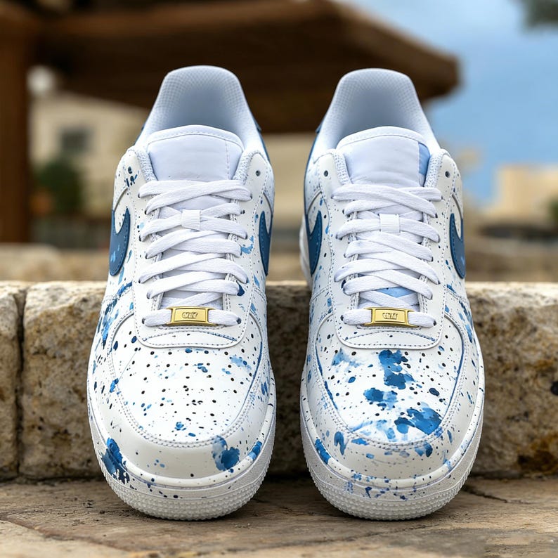 May include: Pair of white sneakers with blue paint splatter design. The shoes have white laces, blue swoosh logos, and gold-colored lace locks. The sneakers are displayed on a stone surface.