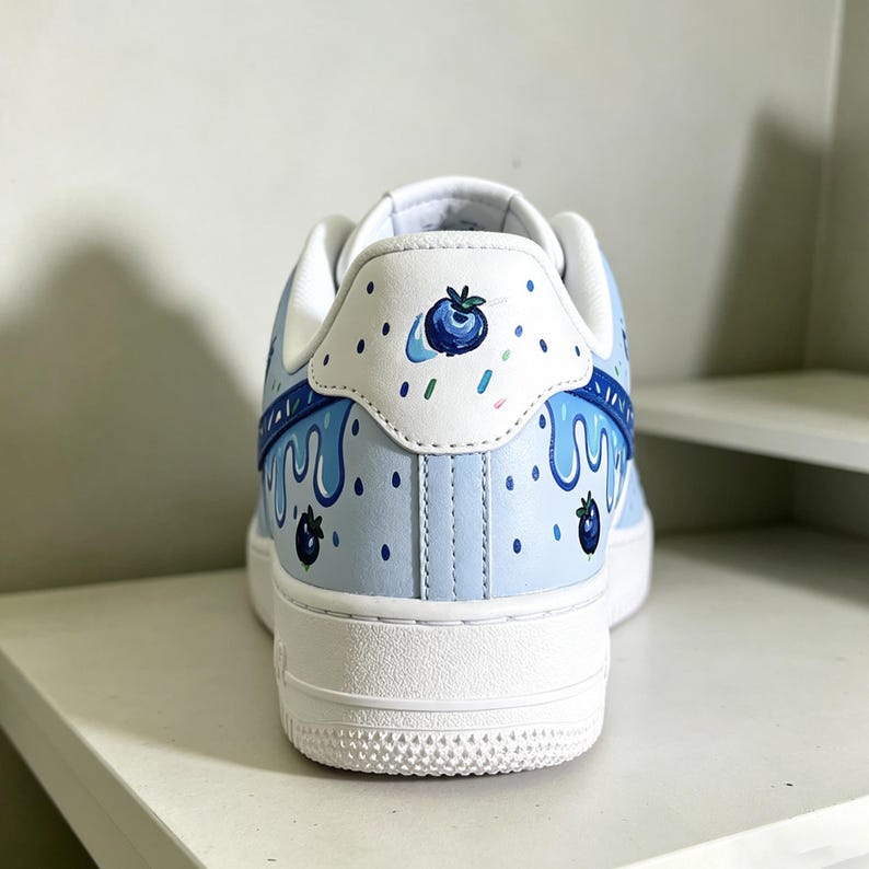 May include: A light blue sneaker with a white sole and heel. The shoe features a blueberry-themed design, with painted blueberries, drips, and sprinkles. The heel has a white patch with a blueberry illustration.