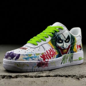May include: White leather sneaker featuring a colorful graffiti design and a portrait of the Joker. The shoe has bright green laces and a white sole. The design includes red, yellow, green, and purple paint splatters.