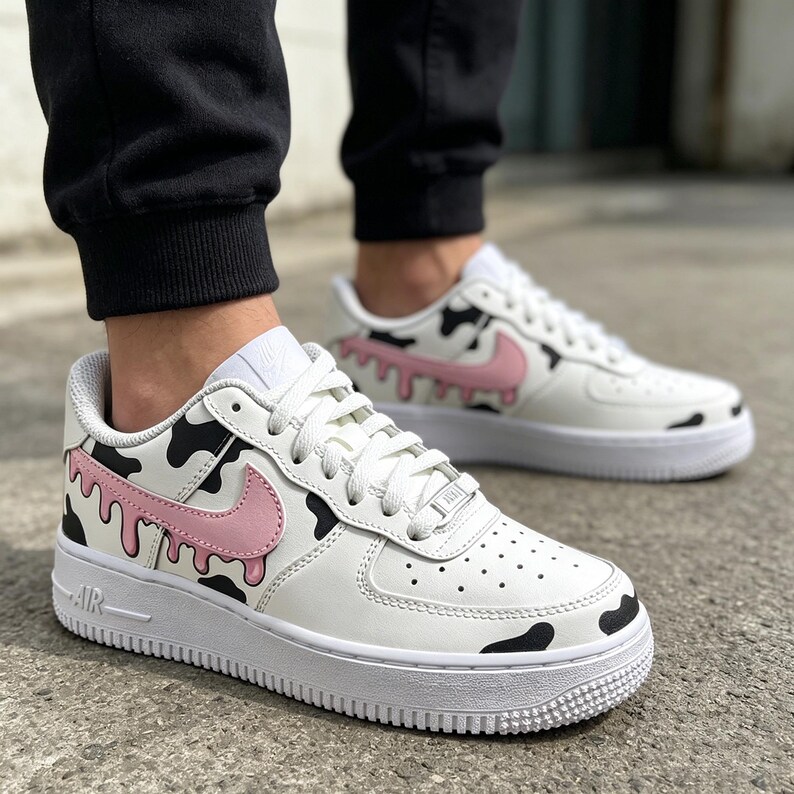 Op de afbeelding: Witte leren sneakers met een uniek koeienprint ontwerp. De schoenen hebben zwarte vlekken, roze swooshes en een roze druppeleffect. De zolen zijn wit en de veters zijn wit.