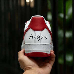 Puede incluir: Zapatilla deportiva blanca y roja con el nombre "Angus" impreso en el tal&oacute;n. La zapatilla presenta una base blanca con detalles rojos en el tal&oacute;n, la suela y los laterales. La zapatilla est&aacute; sujeta por una mano.